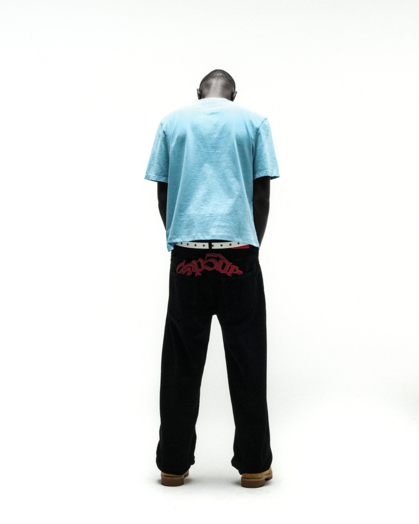 Man from behind wearing a light blue cotton tee and black pants with red embroidered text.
