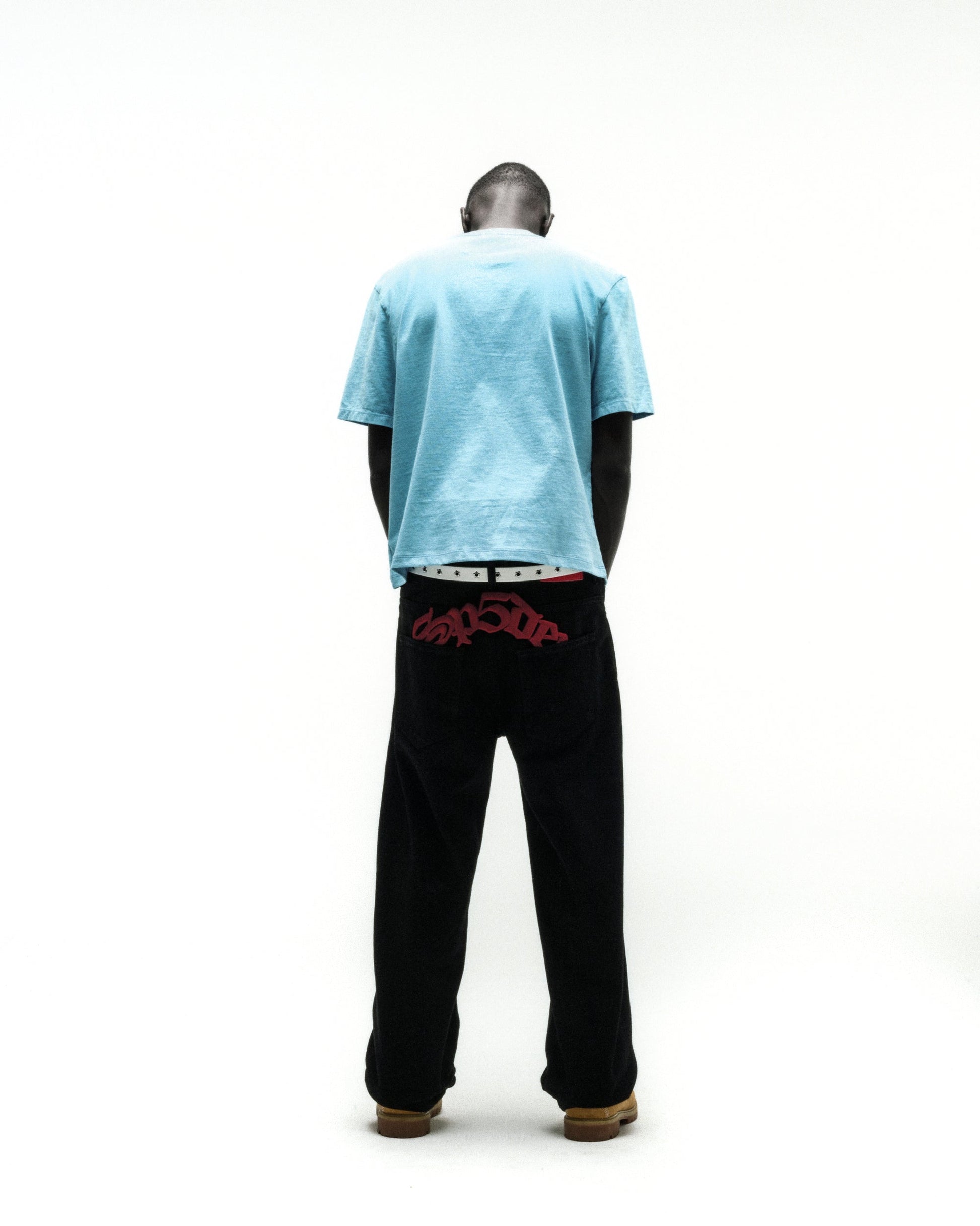 Man from behind wearing a light blue cotton tee and black pants with red embroidered text.