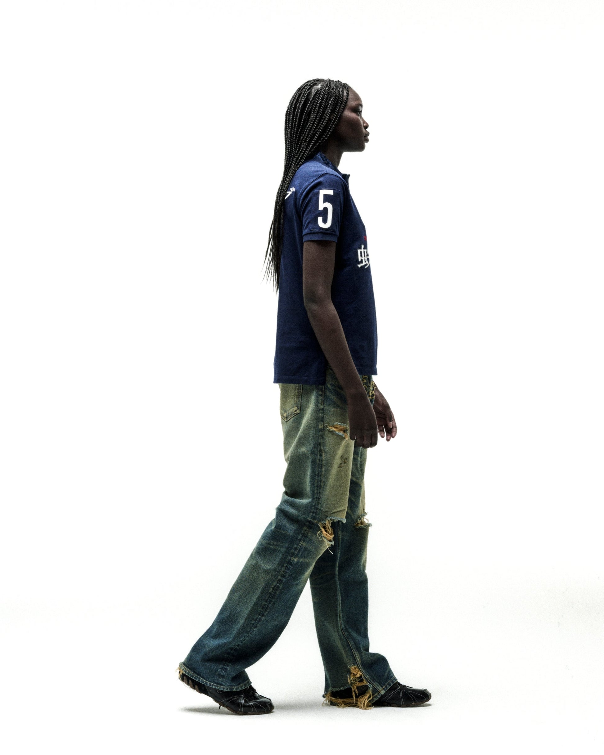 Dark-skinned model in a navy blue polo shirt with "5" and white text, paired with distressed denim jeans.