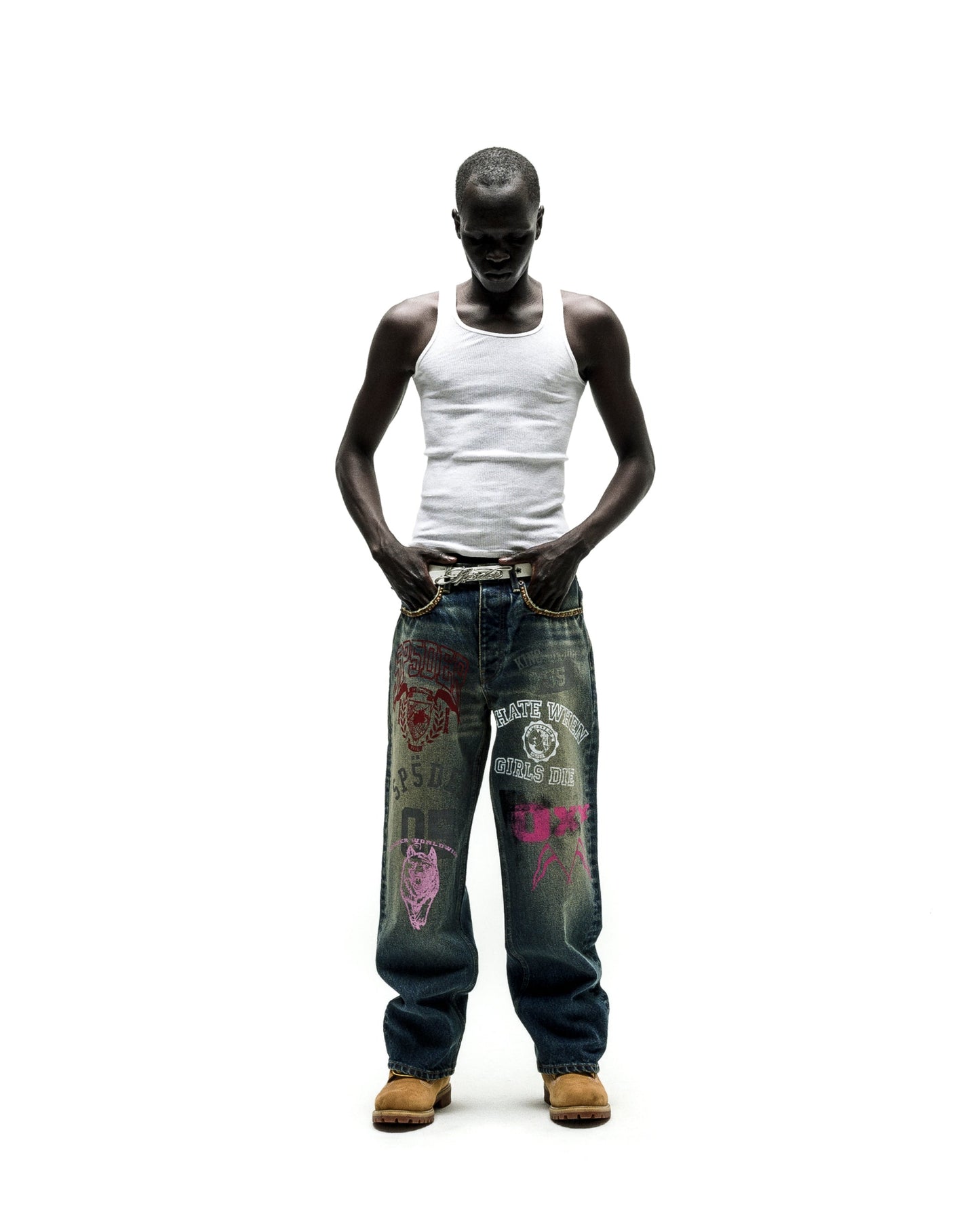 Man in a white tank top, baggy blue jeans with colorful distressed patches, and tan boots.