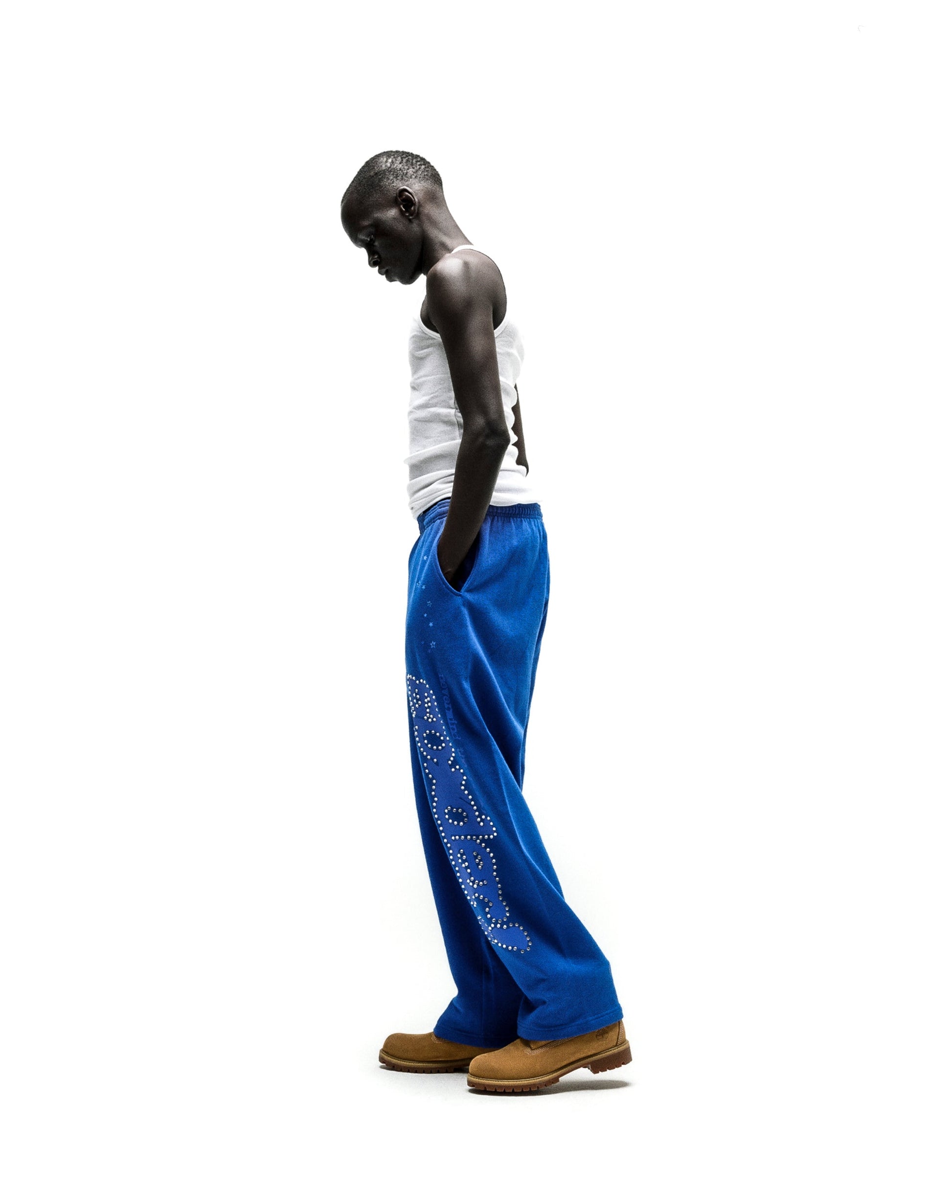 Model in blue RHINESTONE PUNK V2 SWEATPANTS with "P*NK SP5DER" text and scattered stars, white tank top, and tan boots.
