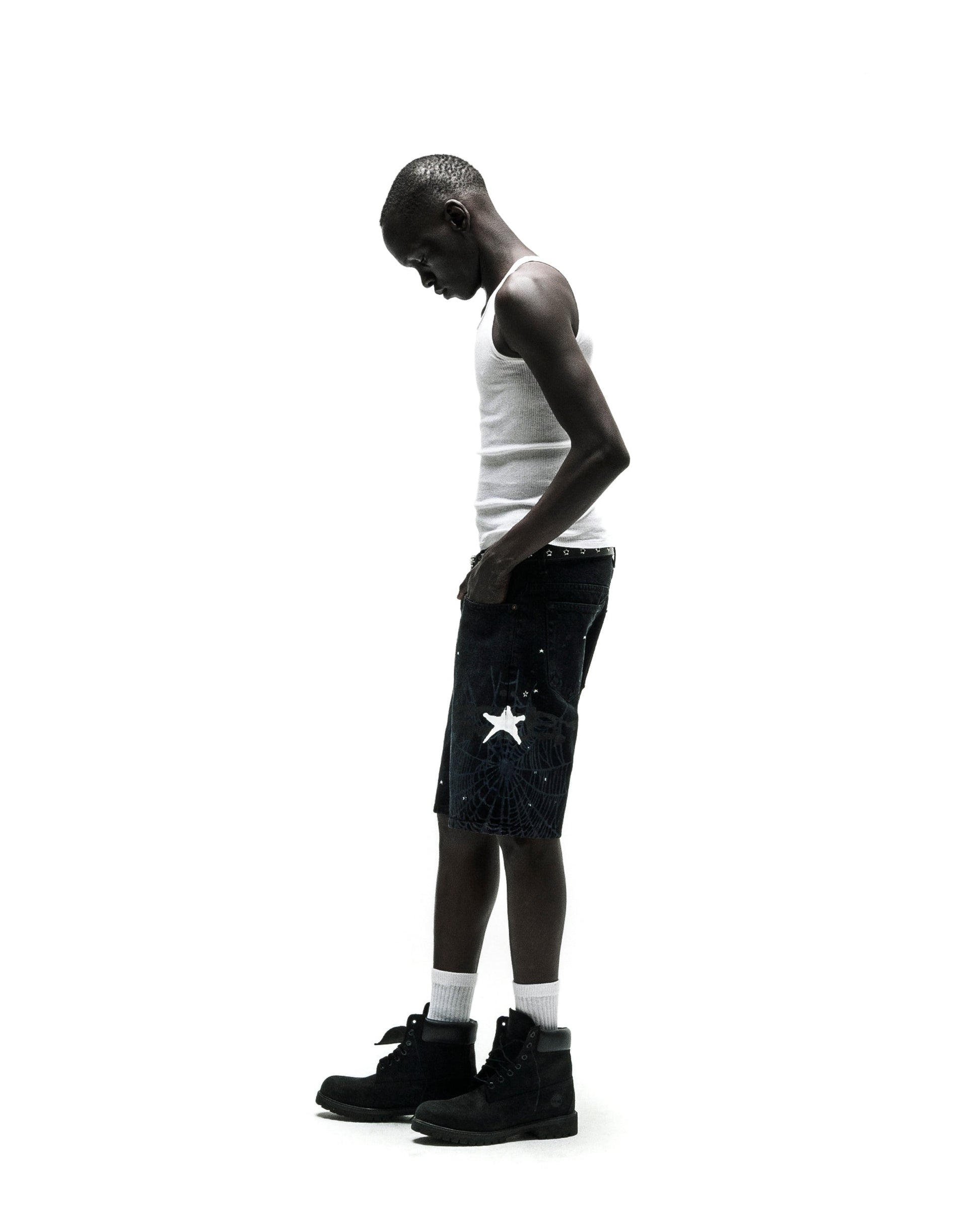 Man in dark wash HEAVY WEB JEAN SHORTS with white star graphic, white tank top, and black boots.