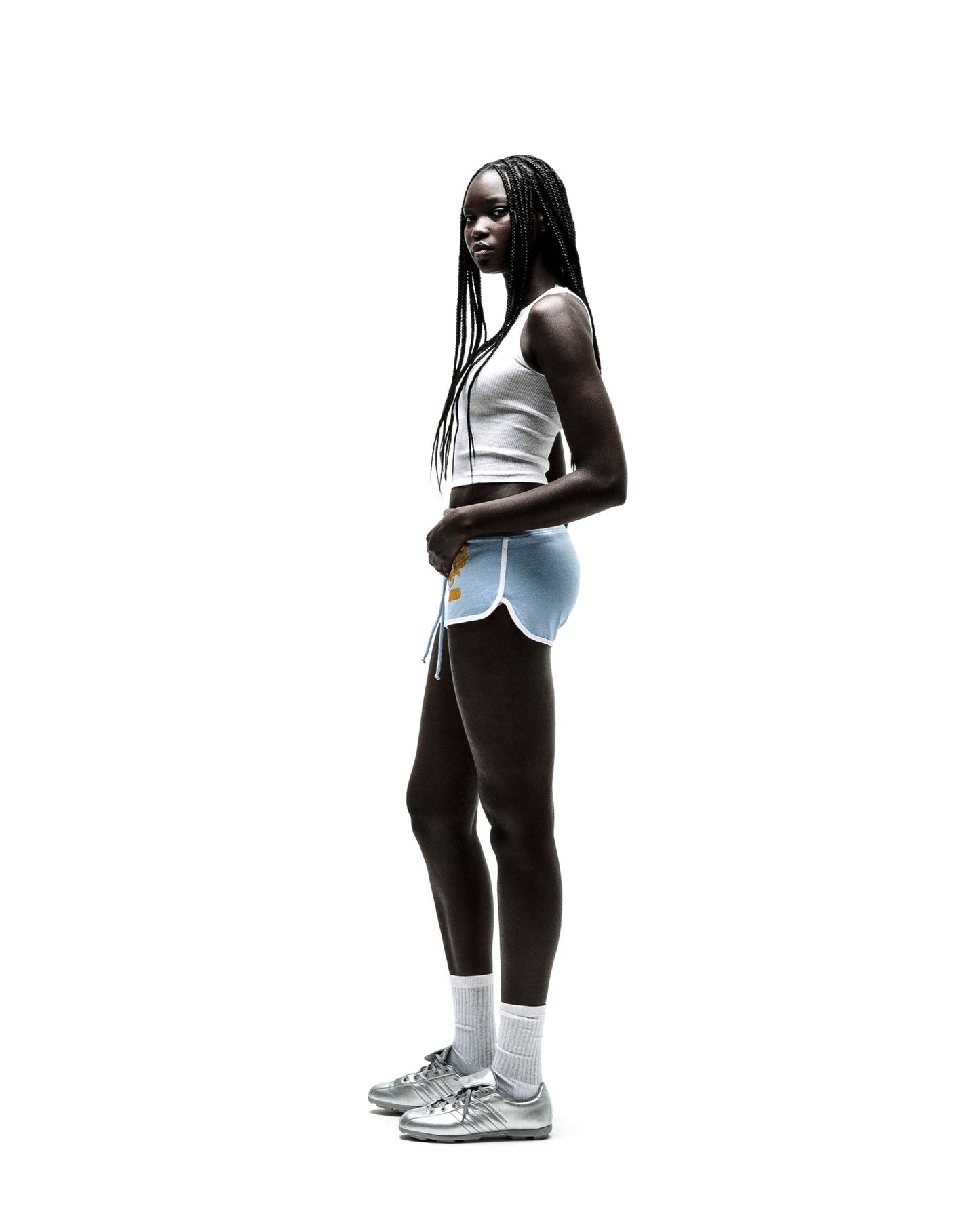Young woman in light blue SP5 WIDE BOXER shorts with white trim, a white crop top, and silver sneakers.