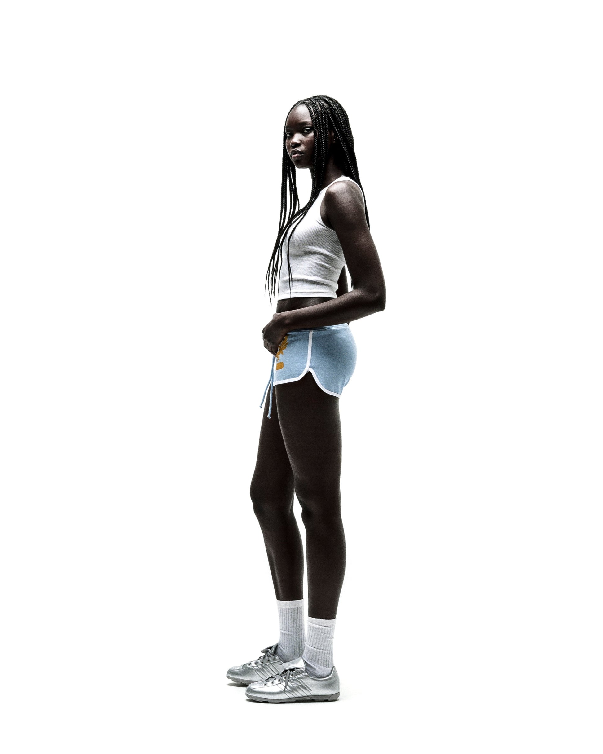 Young woman in light blue SP5 WIDE BOXER shorts with white trim, a white crop top, and silver sneakers.