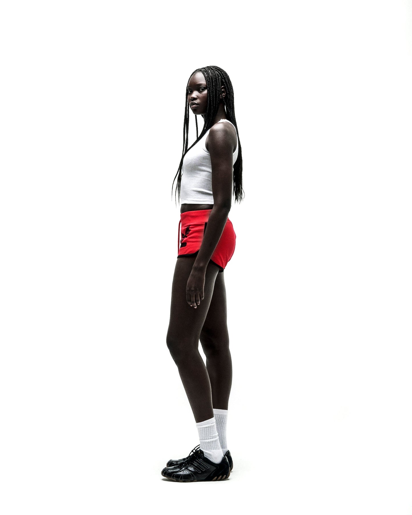 Woman in SP5 WIDE BOXER red shorts with black logo, white tank top, and black sneakers.
