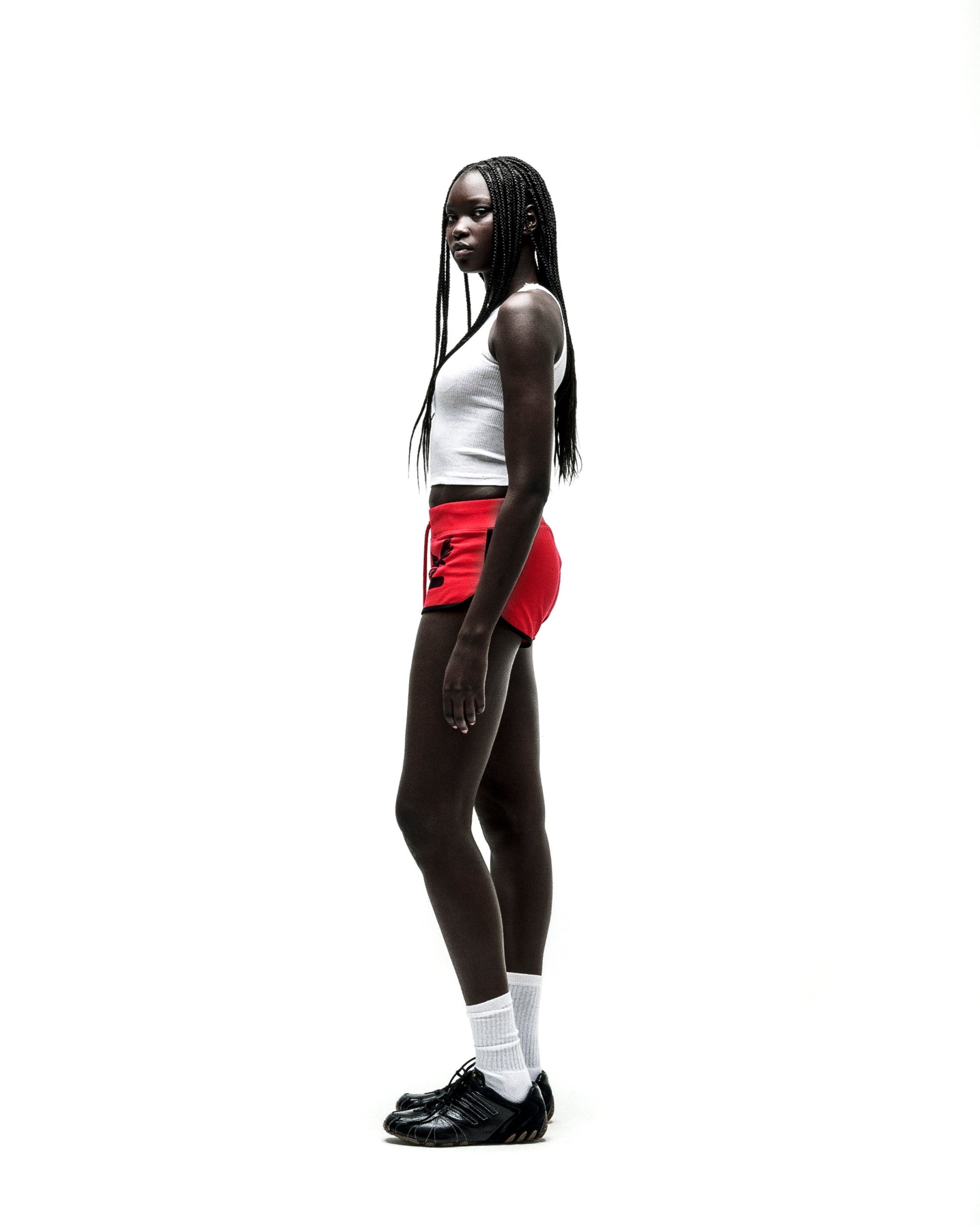 Woman in SP5 WIDE BOXER red shorts with black logo, white tank top, and black sneakers.